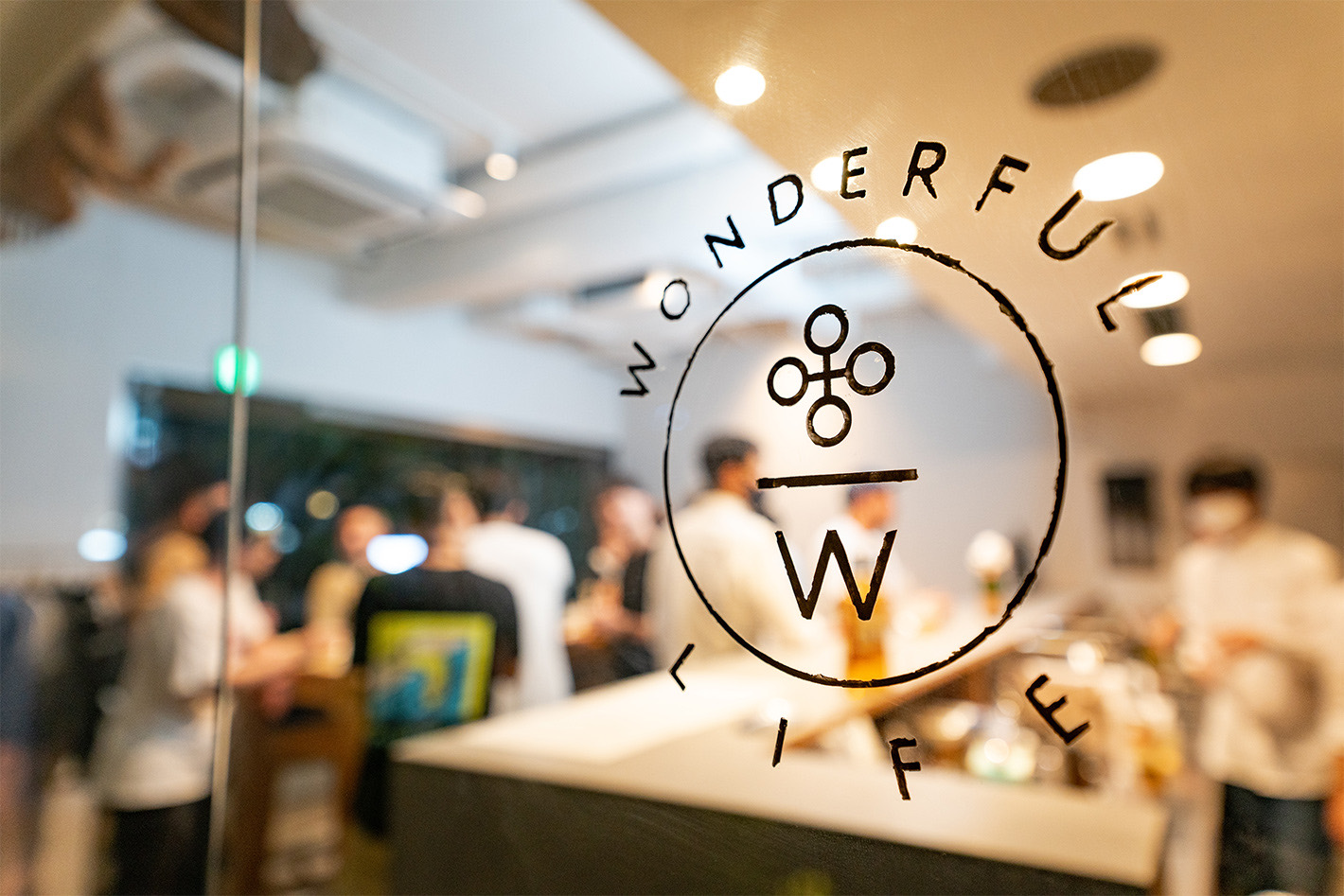 ガラスに描かれた「WONDERFUL LIFE」のロゴマーク。輪の中に四つの小さな輪とW。その奥に、ぼやけた店内と集まった人々。