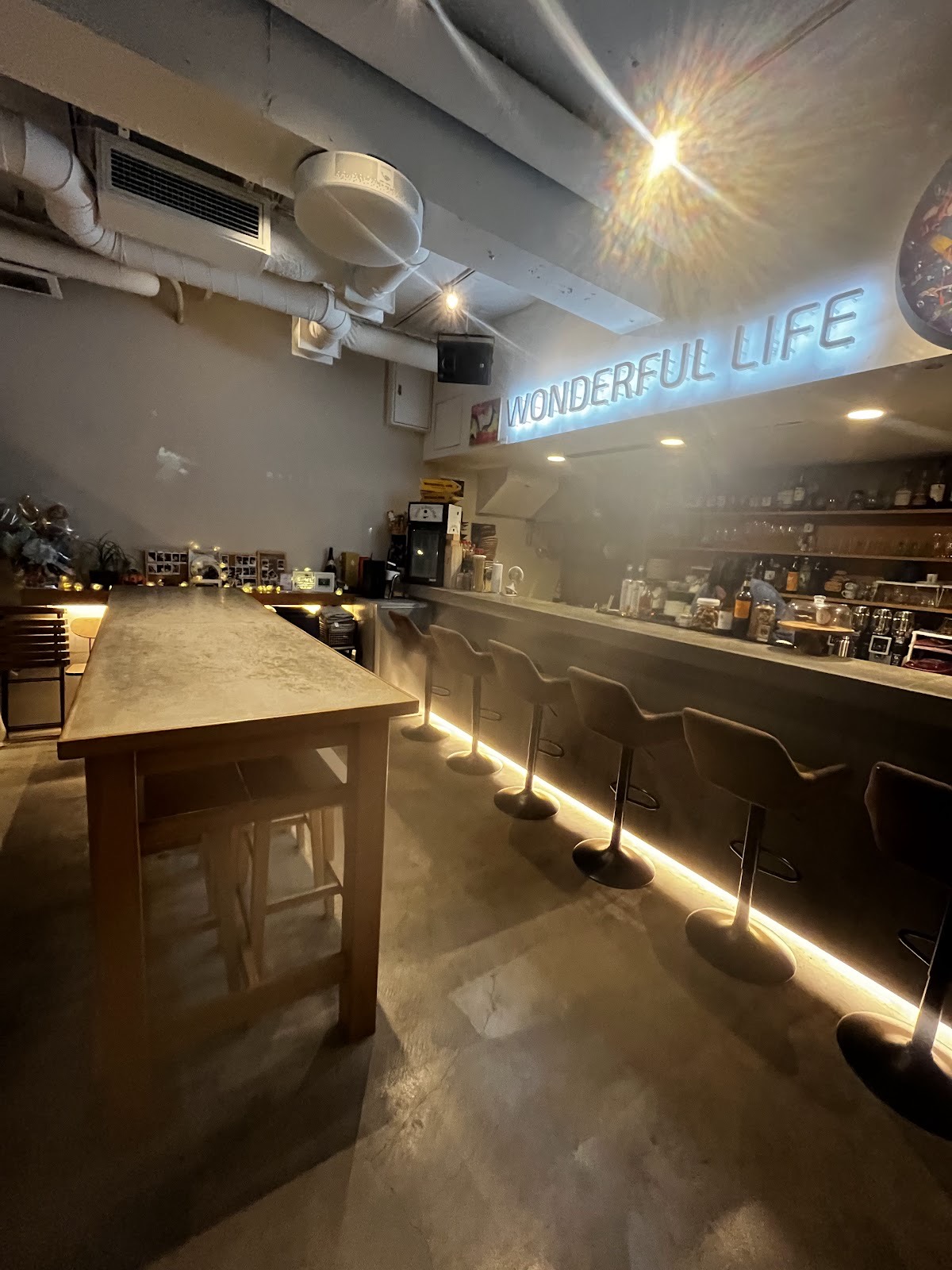 夜の店内。コンクリートのカウンターに並ぶ六脚のスツール、足元の間接照明、奥の棚にボトルとグラス。壁に青白く光る「WONDERFUL LIFE」の文字。左に木の長いハイテーブル。